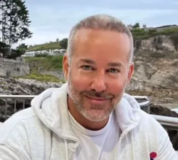 Smiling man with short gray hair and a gray beard, wearing a white hoodie outdoors by a rocky coast